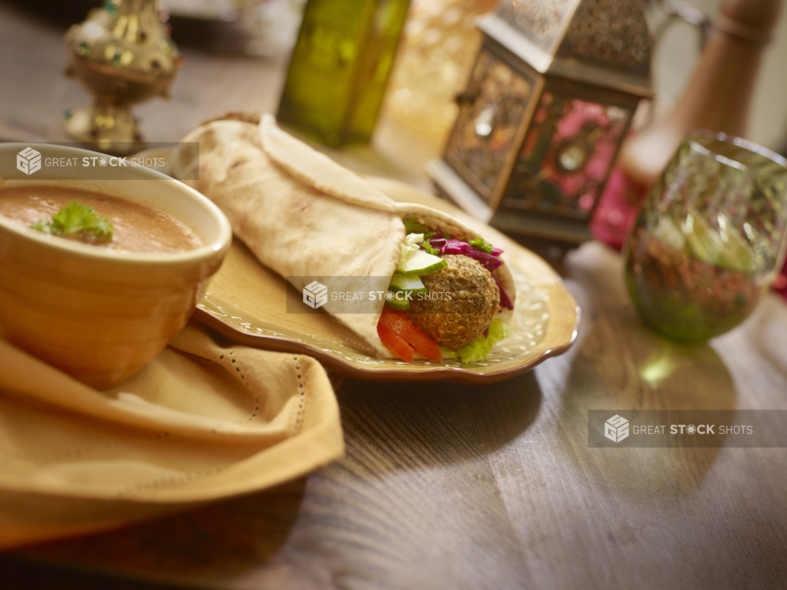 Falafel Pita Wrap with Pickled Veggies and Fresh Vegetables on a Coloured Ceramic Dish with a Bowl of Thick Hot Vegetarian Soup on a Wooden Table in an Indoor Setting