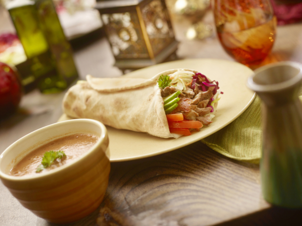 Chicken Shawarma Pita Wrap with Pickled Veggies and Fresh Vegetables on a Coloured Ceramic Dish with a Bowl of Thick Hot Vegetarian Soup on a Wooden Table in an Indoor Setting
