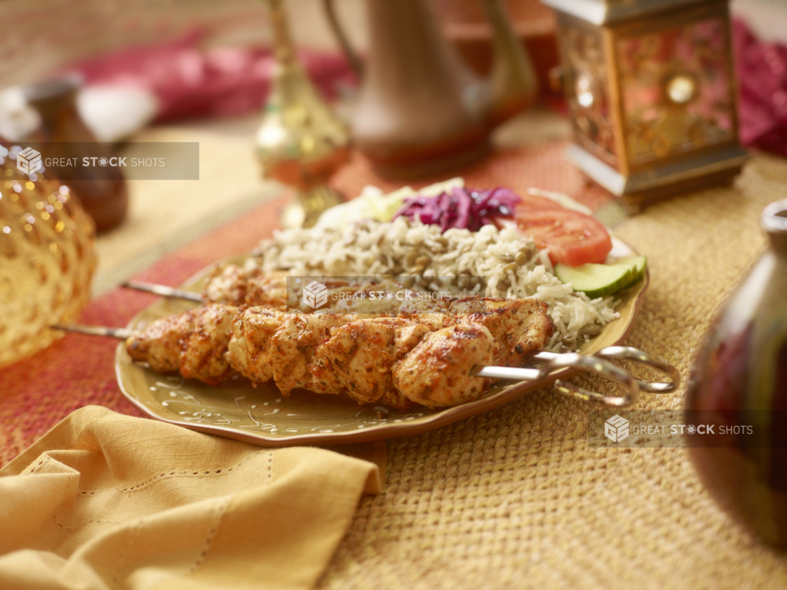 Combo Meal of BBQ Chicken Kebab on Metal Skewers with a Garden Salad, Picked Veggies, Rice and Lentils in a Ceramic Dish on a Woven Placemat in an Indoor Setting