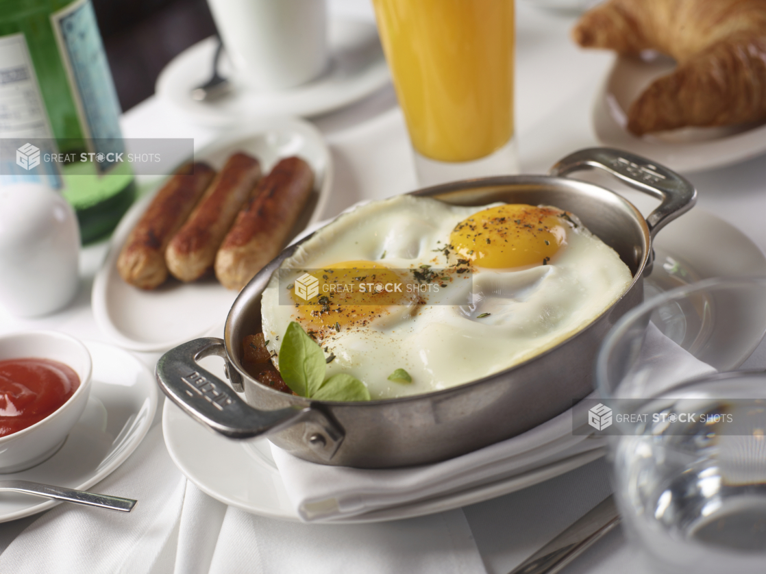 Two Sunny Side Up Fried Eggs and Breakfast Potatoes in an Individual Skillet with Sausages and Other Breakfast Items on a White Table Cloth Table Setting