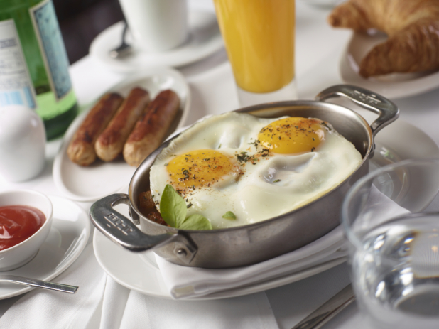 Two Sunny Side Up Fried Eggs and Breakfast Potatoes in an Individual Skillet with Sausages and Other Breakfast Items on a White Table Cloth Table Setting
