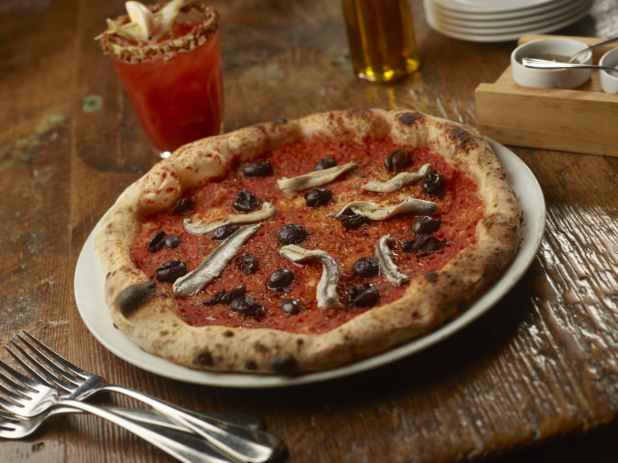 Neopolitan-style Pizza with Anchovies and Mediterranean Olives and Marinara sauce on a Round White Plate on a Wooden Surface with a Blood Caesar Cocktail in the Background