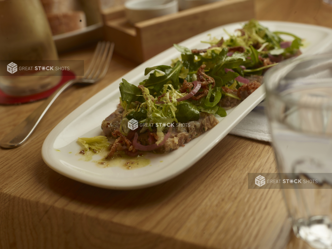 A Starter of Tuna Tartare Topped with Baby Arugula, Curled Endive, Pickled Red Onions and Crispy Fried Onions on a White Ceramic Platter in a Restaurant Setting