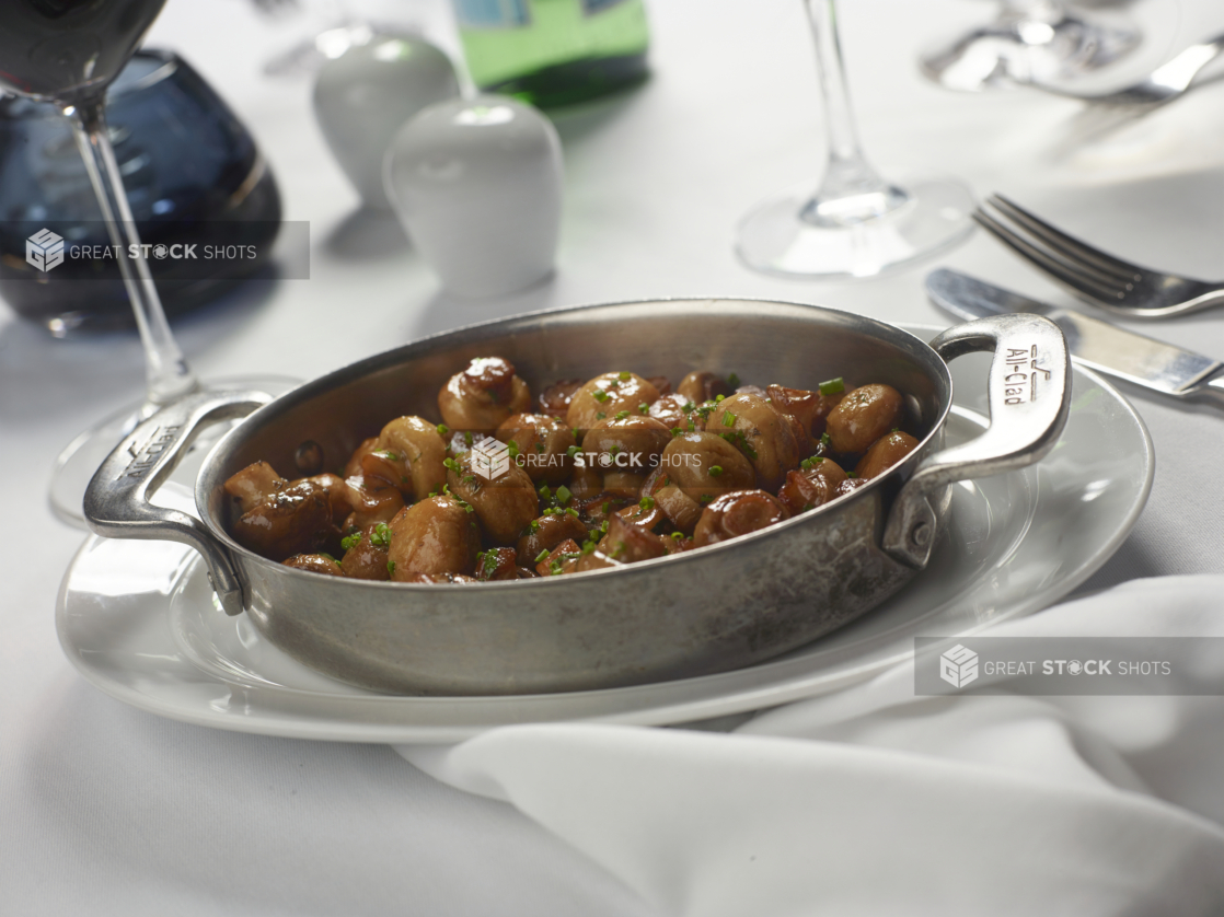 Large Stainless Steel Skillet with Sautéed Whole Mushrooms in a Brown Sauce with Chive Garnish on a White Table Cloth Table Setting