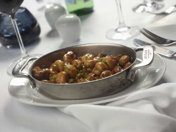 Large Stainless Steel Skillet with Sautéed Whole Mushrooms in a Brown Sauce with Chive Garnish on a White Table Cloth Table Setting