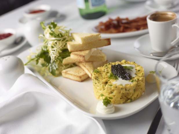A Fancy Platter of Scrambled Eggs with Caviar and Parsley with Frisée and Crustless Toast Points and other Breakfast Items on a White Table Cloth Table Setting