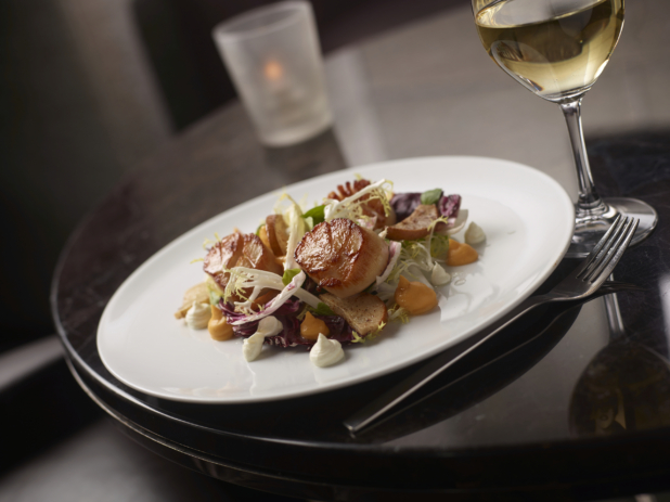 Seared Scallops and Roasted Pear on a Bed of Frisée and Radicchio Salad with a Glass of White Wine on a Round Dark Wood Table in a Restaurant Setting