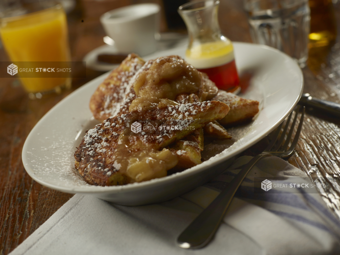 French Toast with Apple Cinnamon Topping and Dusted with Powdered Sugar on a Round White Dish with Other Breakfast Items on a Wooden Table in a Casual Dining Setting