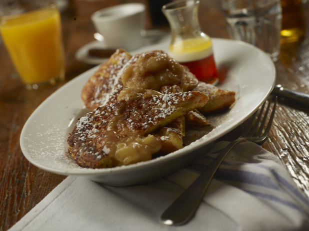 French Toast with Apple Cinnamon Topping and Dusted with Powdered Sugar on a Round White Dish with Other Breakfast Items on a Wooden Table in a Casual Dining Setting