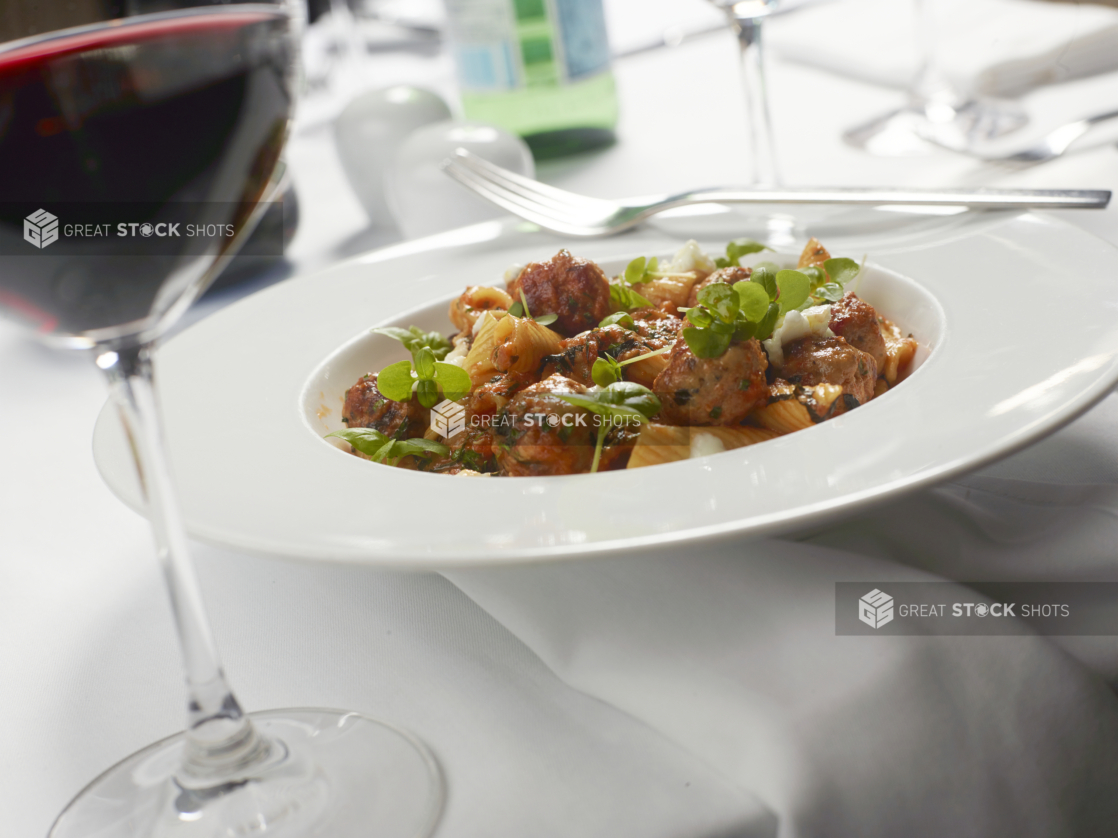 Rigatoni and Herbed Meatballs in a Marinara Sauce with Fresh Cheese in a Round White Bowl with a Glass of Red Wine on a White Table Cloth Table Setting