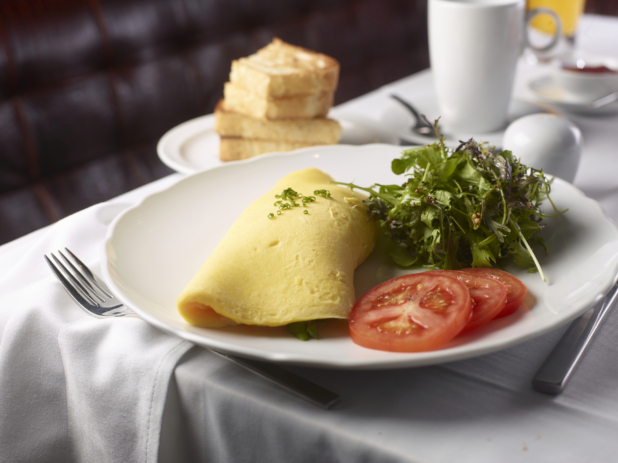 A Plain Omelette with a side of Spring Mix Salad and Sliced Tomatoes on a Round White Plate with Toast and Other Breakfast Items on a White Table Cloth Table Setting
