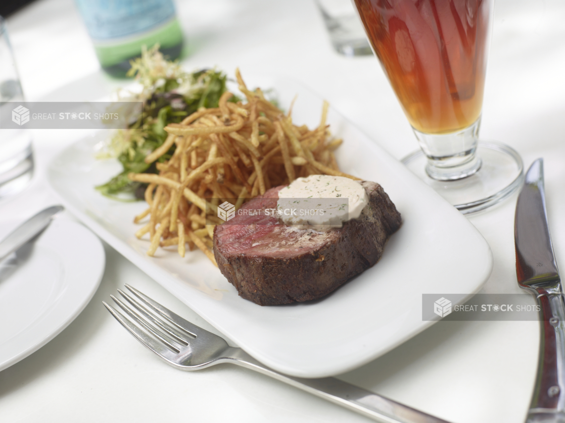 Steak Frites with a Pat of Herbed Butter and a Side of Mixed Greens Salad on a White Platter with a Glass of Dark Beer on a White Table Cloth Outdoor Table Setting