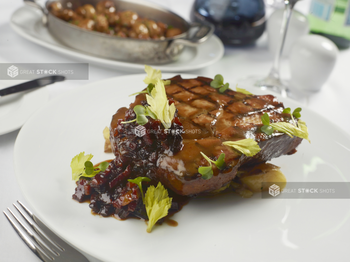 Grilled Pork Steak with Brown Butter Glaze and Duck Fat Balsamic Reduction over Potato Purée on a Round White Plate in a Fine Dining Table Setting