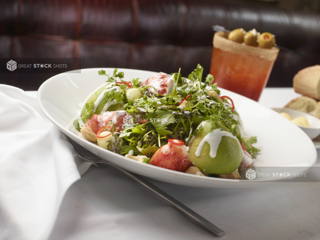 Seafood Salad with Lobster Meat and Avocado on a Bed of Mixed Greens on a Round White Dish with a Bloody Caesar, Bread and Butter on a White Table Cloth Table Setting