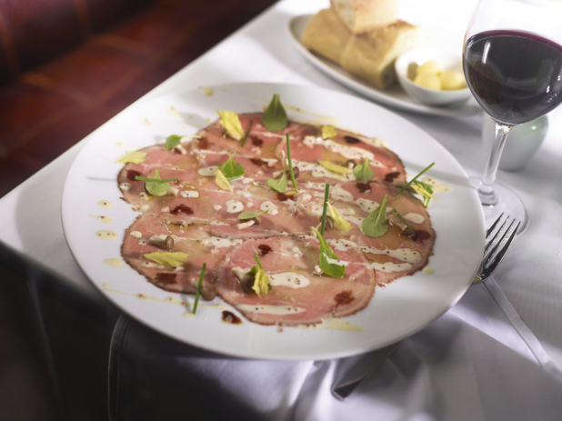 Carpaccio with Aioli Dressing and Mixed Greens on a Round White Plate with a Glass of Red Wine and a Platter of Bread and Butter on a White Table Cloth Table Setting