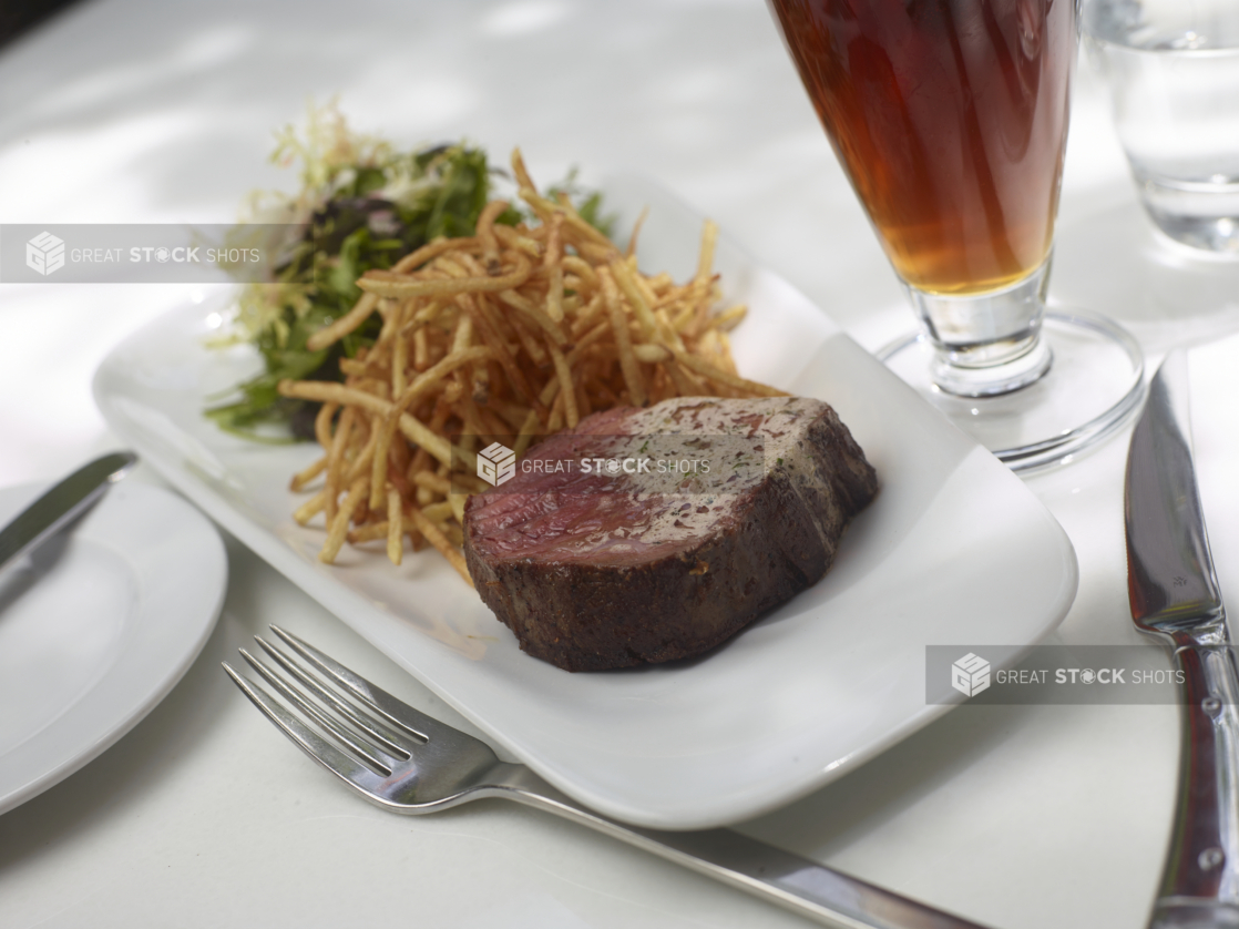 Steak Frites with a Side of Mixed Greens Salad on a White Platter with a Glass of Dark Beer on a White Table Cloth Outdoor Table Setting