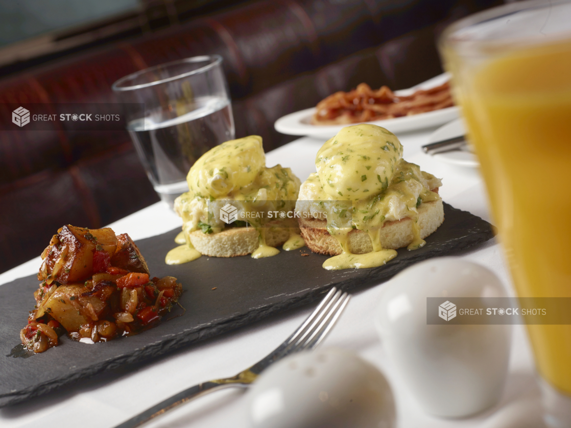 Eggs Benedict with a Side of Roasted Potatoes and Vegetables and Other Breakfast Items on a White Table Cloth Table Setting