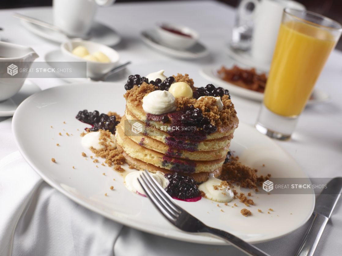 A Stack of Fluffy Pancakes with Blueberry Coulis and Dollops of Cream and Butter on a Round White Plate and other Breakfast Items on a White Table Cloth Covered Dining Table