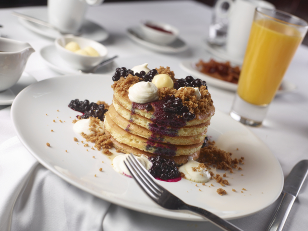 A Stack of Fluffy Pancakes with Blueberry Coulis and Dollops of Cream and Butter on a Round White Plate and other Breakfast Items on a White Table Cloth Covered Dining Table