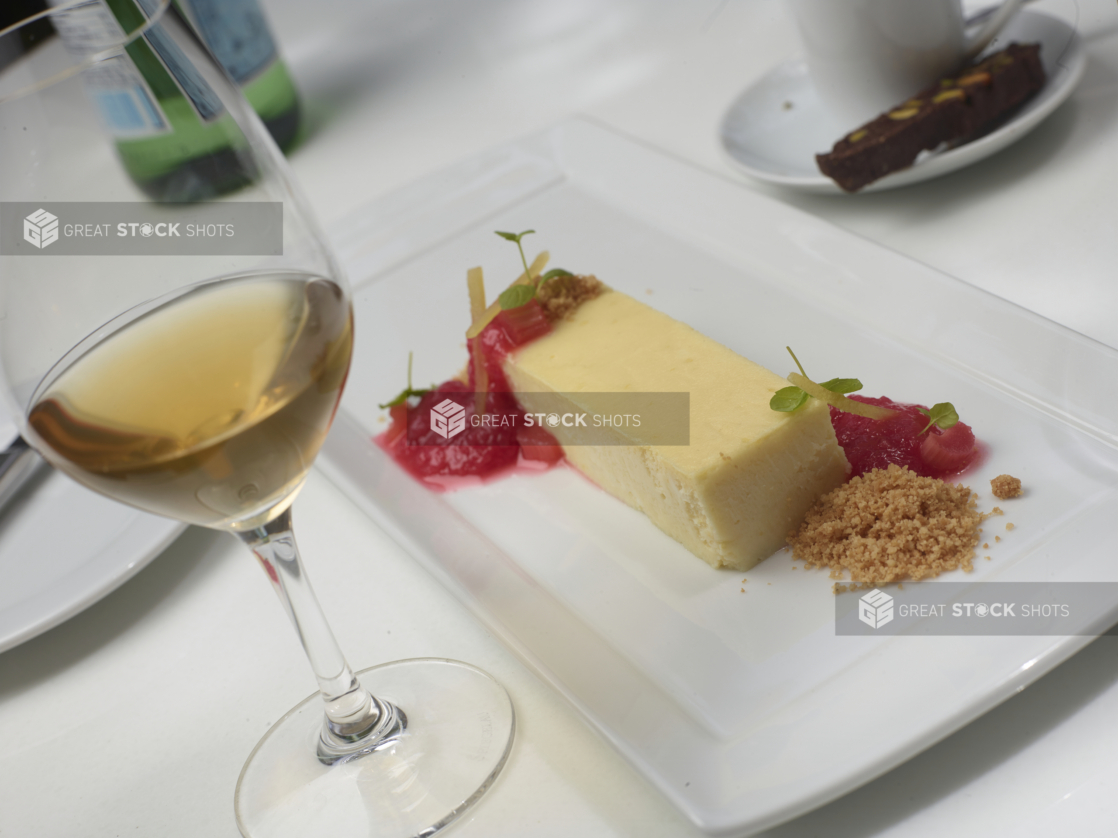 Cheesecake with Strawberry and Rhubarb Coulis and Graham Cracker Crumble on a White Rectangular Platter with a Glass of White Wine, Coffee and Biscotti on a White Table Cloth in an Outdoor Setting