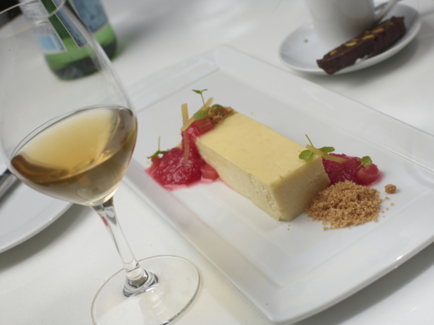 Cheesecake with Strawberry and Rhubarb Coulis and Graham Cracker Crumble on a White Rectangular Platter with a Glass of White Wine, Coffee and Biscotti on a White Table Cloth in an Outdoor Setting