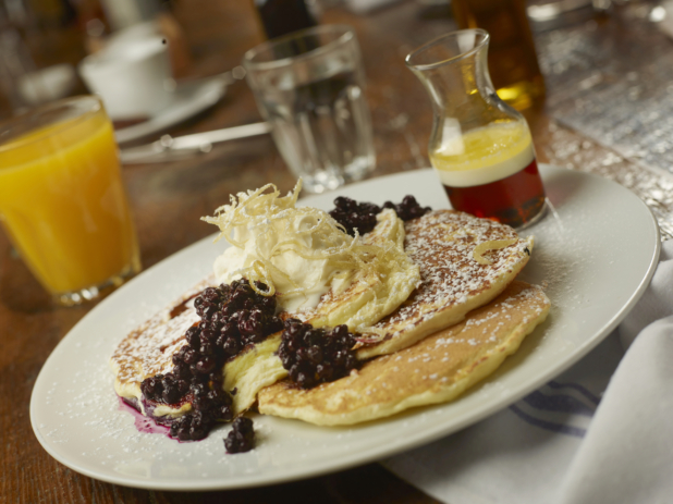 Pancakes with Whipped Cream and Blueberry Coulis Dusted with Powdered Sugar on a Round White Dish with Other Breakfast Items on a Wooden Table in a Casual Dining Setting