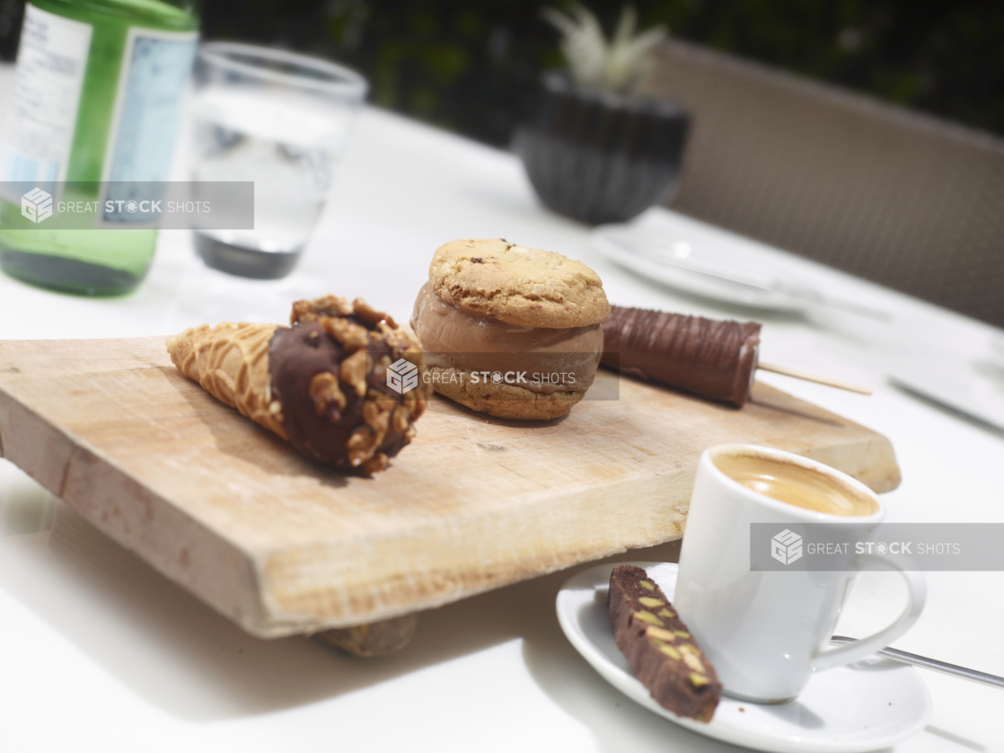 A Trio of Mini Frozen Desserts - Ice Cream Sandwich, Chocolate-Dipped Ice Cream Cone and Fudgesicle Ice Pop on a Wooden Platter with Coffee and Biscotti on a White Table Cloth in an Outdoor Setting