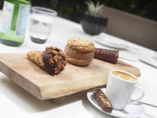 A Trio of Mini Frozen Desserts - Ice Cream Sandwich, Chocolate-Dipped Ice Cream Cone and Fudgesicle Ice Pop on a Wooden Platter with Coffee and Biscotti on a White Table Cloth in an Outdoor Setting