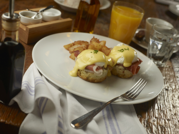 Eggs Benedict and Breakfast Potatoes on a Round White Dish with Other Breakfast Items on a Wooden Table in a Casual Dining Setting