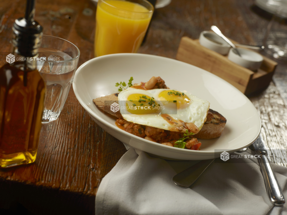 Two Sunny Side Up Fried Eggs Over White Beans in Tomato Vegetable Sauce on a Grilled Sliced Baguette in a Shallow Round White Bowl on a Wooden Table with Other Breakfast Items in a Casual Table Setting