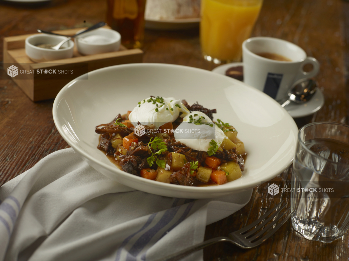 Short Rib Hash in a Thick Brown Sauce with Cubed Vegetables and Two Poached Eggs in a Round White Bowl with Other Breakfast Items on a Wooden Table in a Casual Dining Setting