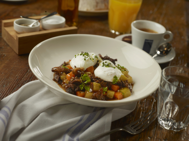 Short Rib Hash in a Thick Brown Sauce with Cubed Vegetables and Two Poached Eggs in a Round White Bowl with Other Breakfast Items on a Wooden Table in a Casual Dining Setting
