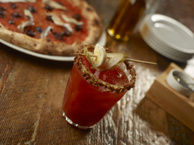 Bloody Caesar with pickled egg and pickle slice garnish and celery salt rim on a wooden table with a neopolitain-style pizza in the background