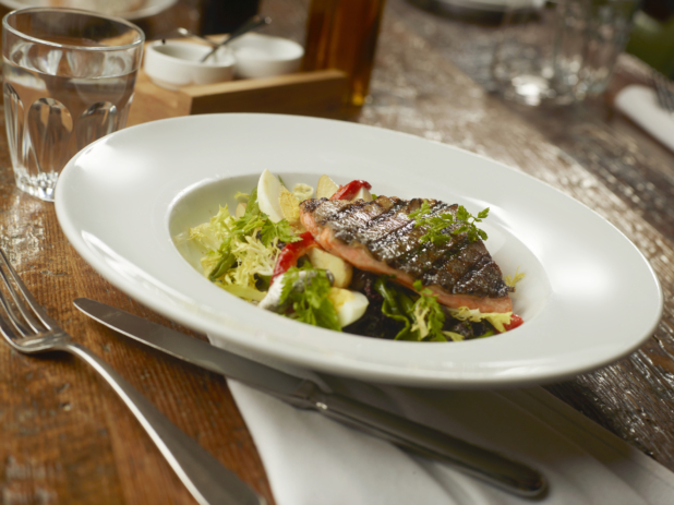 Grilled Salmon Steak on a Bed of Frisée Salad with Sliced Boiled Eggs and Red Pepper in a Round White Shallow Bowl on a Wooden Table in a Restaurant Dining Setting