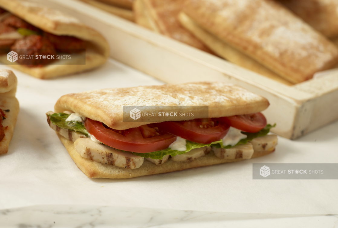 Grilled Chicken Strips, Sliced Tomatoes and Romaine Lettuce in a Fold-over Ciabatta Bun on a Marble Surface with a Meatball Sandwich and Fresh Bread in the Background