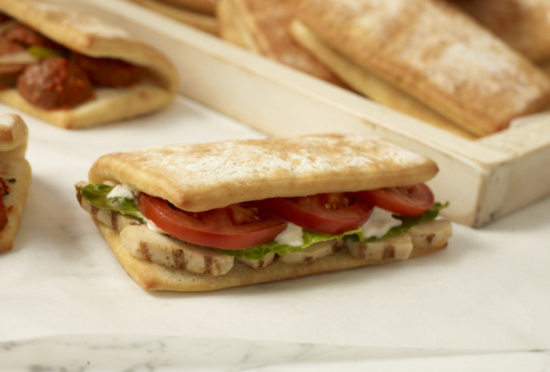 Grilled Chicken Strips, Sliced Tomatoes and Romaine Lettuce in a Fold-over Ciabatta Bun on a Marble Surface with a Meatball Sandwich and Fresh Bread in the Background