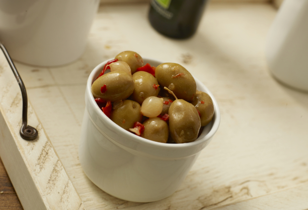 Deep White Ceramic Bowl of Spicy Green Olives with Hot Pepper Flakes and Pearl Onions on a White Painted Wooden Surface