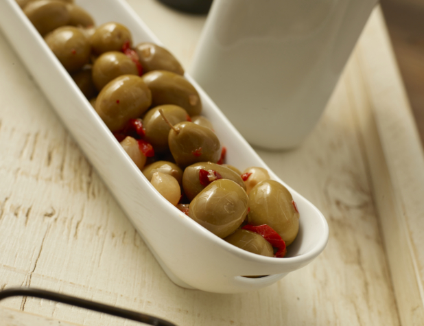 Long White Ceramic Container of Spicy Green Olives with Hot Pepper Flakes and Pearl Onions on a White Painted Wooden Surface
