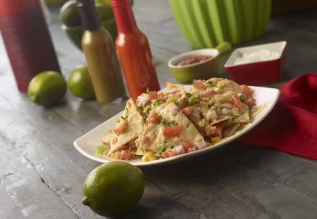 Square White Dish of Nachos and Salsa on a Dark Wood Surface with Bottles of Hot Sauce, Whole Limes and a Red Napkin