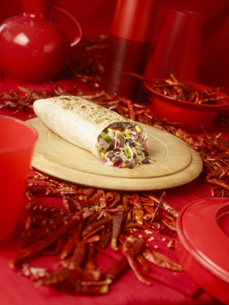 Steak and Bean Burrito Cut Open on a Wooden Platter with Dried Red Chilli Peppers and Red Kitchenware on a Red Placemat