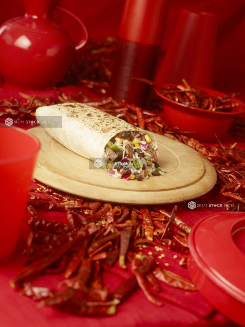 Steak and Bean Burrito Cut Open on a Wooden Platter with Dried Red Chilli Peppers and Red Kitchenware on a Red Placemat