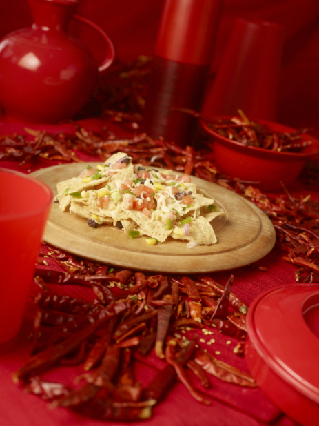 Nacho Chips and Toppings on a Wooden Platter with Dried Red Chilli Peppers and Red Kitchenware on a Red Placemat