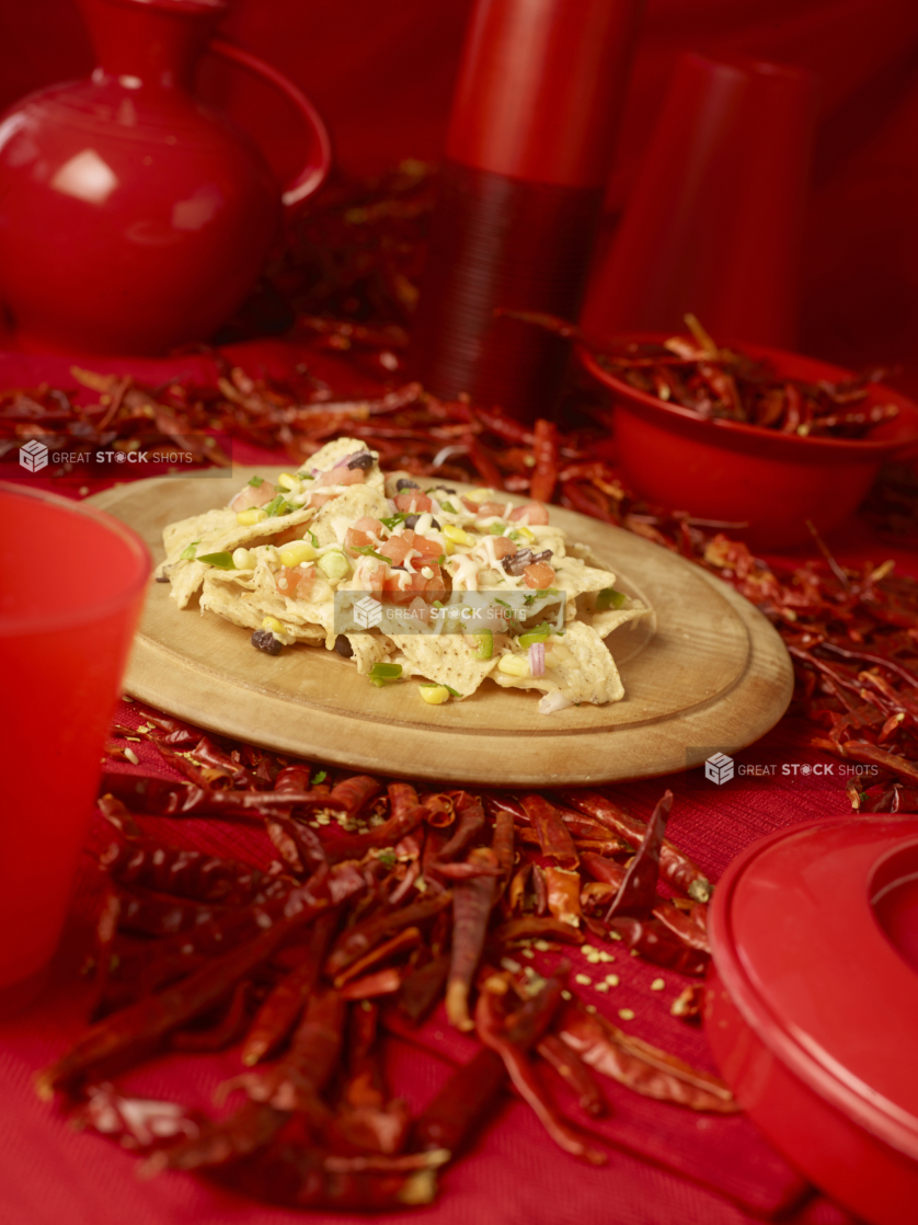 Nacho Chips and Toppings on a Wooden Platter with Dried Red Chilli Peppers and Red Kitchenware on a Red Placemat