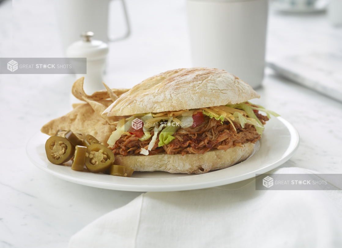 Pulled pork sandwich on a ciabatta bun, with pickled jalapenos and tortilla chips on the side, white table setting