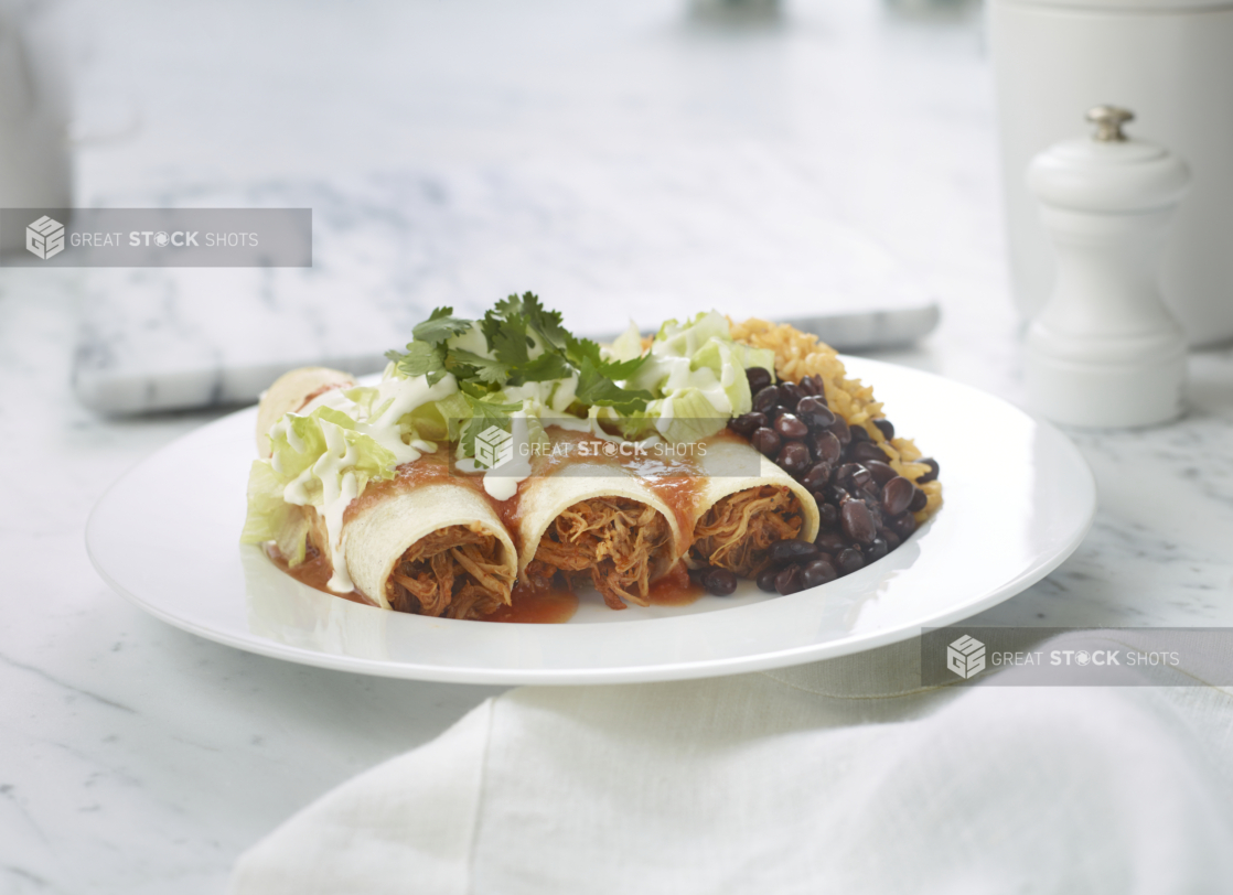 Meat enchiladas with black beans and rice on a white plate on a white marble background