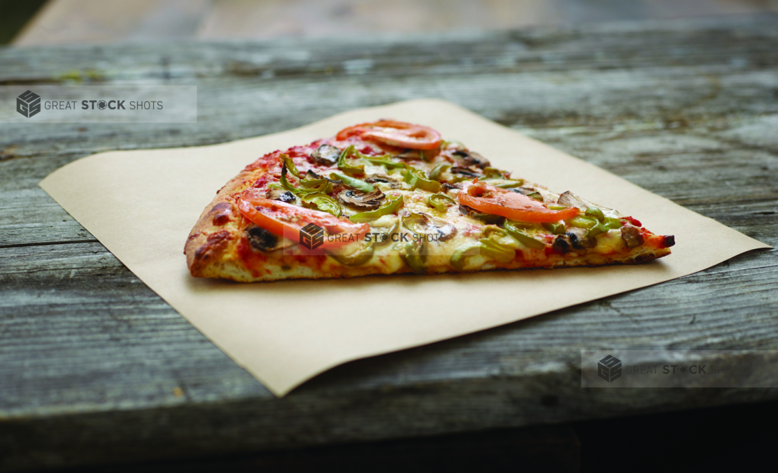 A Jumbo Slice of Roasted Veggie Pizza with Tomatoes, Green Peppers and Mushrooms on Parchment Paper on an Aged Wooden Background