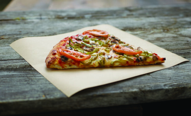 A Jumbo Slice of Roasted Veggie Pizza with Tomatoes, Green Peppers and Mushrooms on Parchment Paper on an Aged Wooden Background