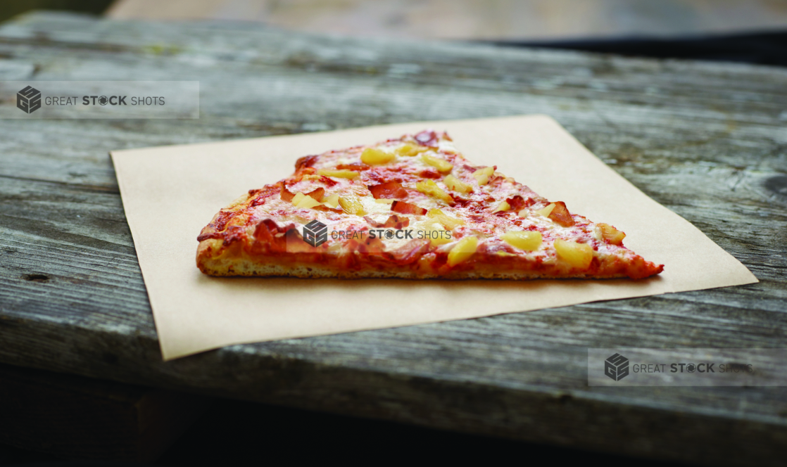 A Jumbo Slice of Hawaiian Pizza on Parchment Paper on an Aged Wood Background