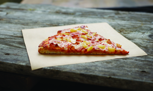 A Jumbo Slice of Hawaiian Pizza on Parchment Paper on an Aged Wood Background