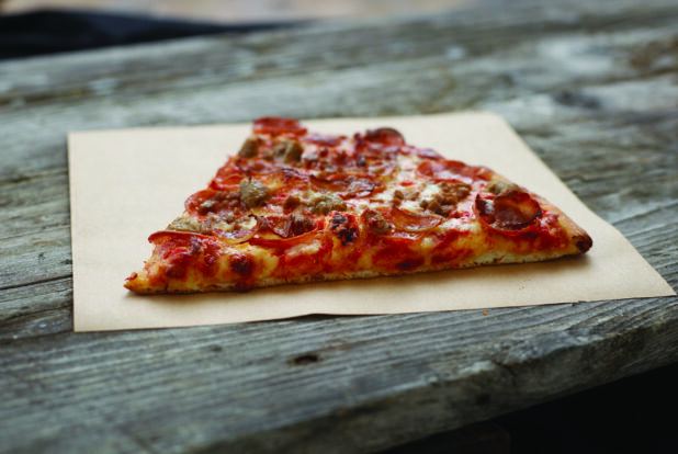 A Jumbo Slice of Meat Lovers Pizza with Pepperoni, Sausage, Bacon and Ham on Parchment Paper on an Aged Wood Background